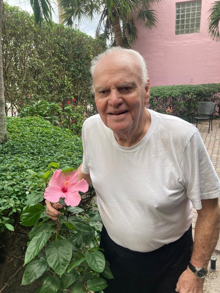 An elderly man smiling while holding a pink flower in a garden.