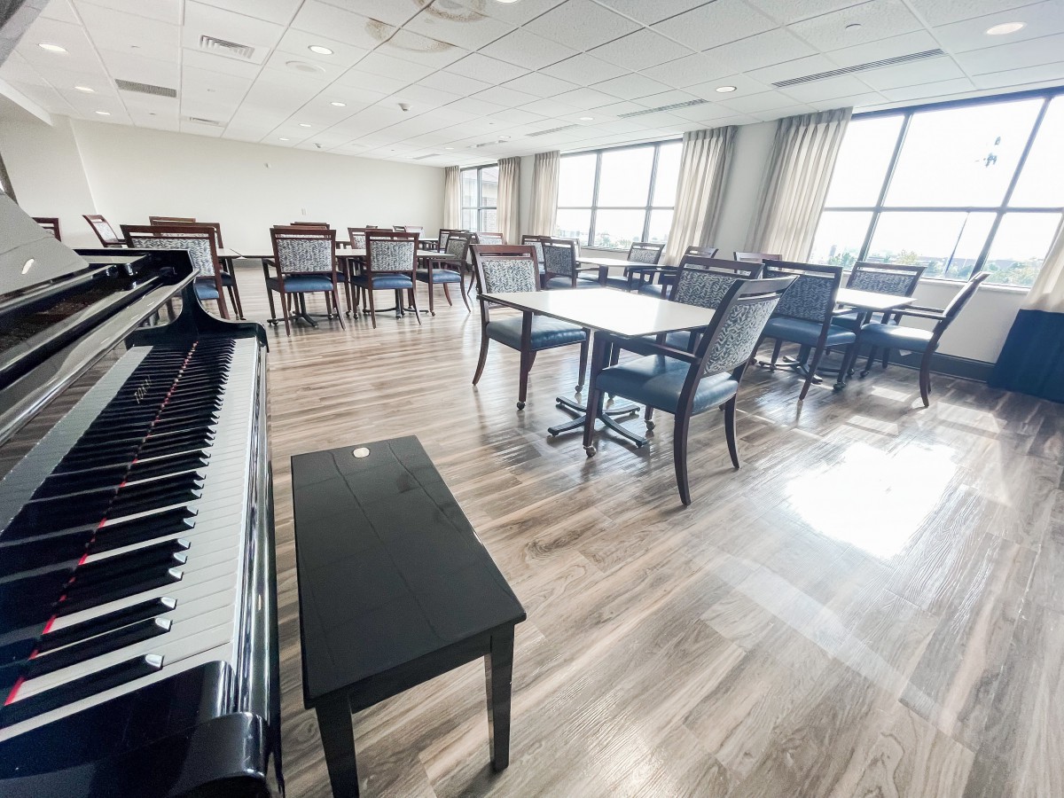 A bright room with a piano, tables, and chairs arranged on a hardwood floor.
