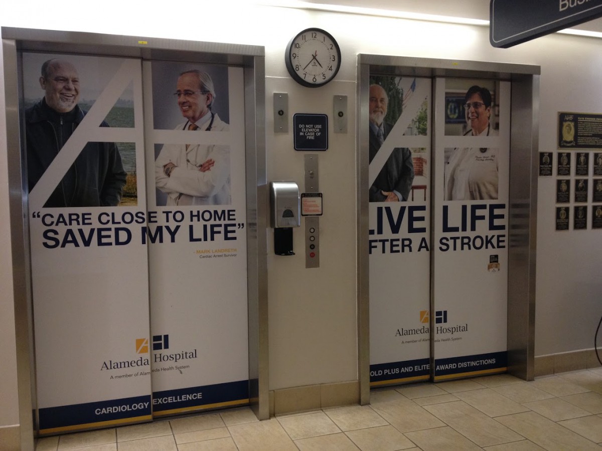 Elevator doors in a hospital featuring healthcare advertisements and a clock above.