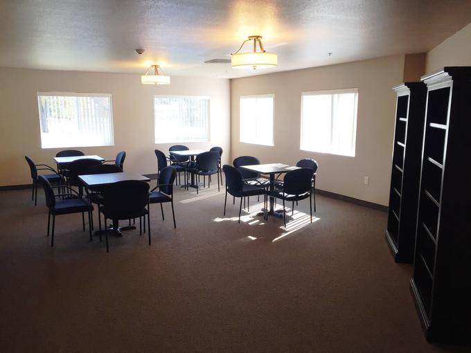A well-lit room with multiple tables and chairs, featuring large windows and carpeted flooring.