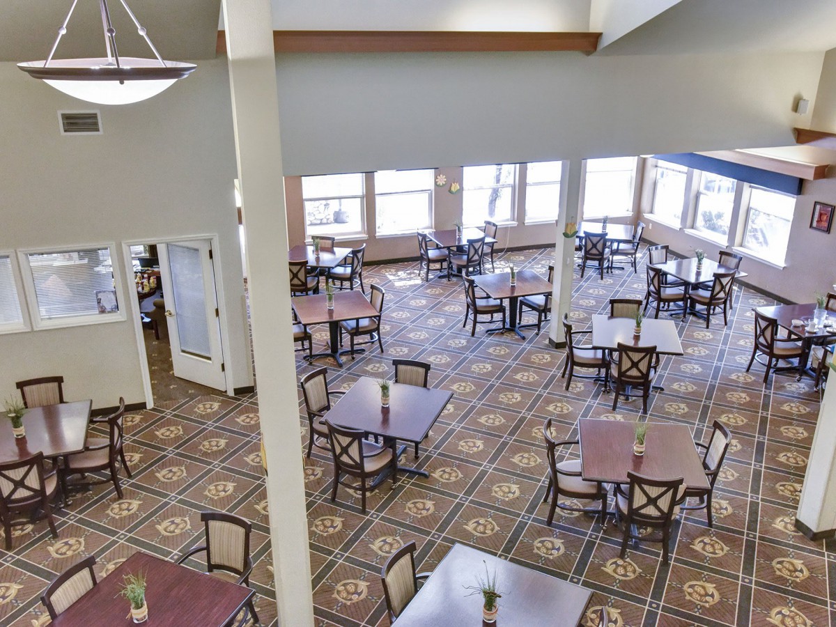 A spacious dining area with multiple tables and chairs, featuring large windows and carpeted flooring.