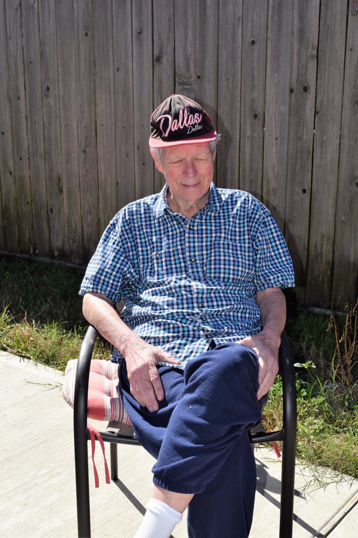 A man wearing a hat and checkered shirt sits outdoors on a chair by a fence.