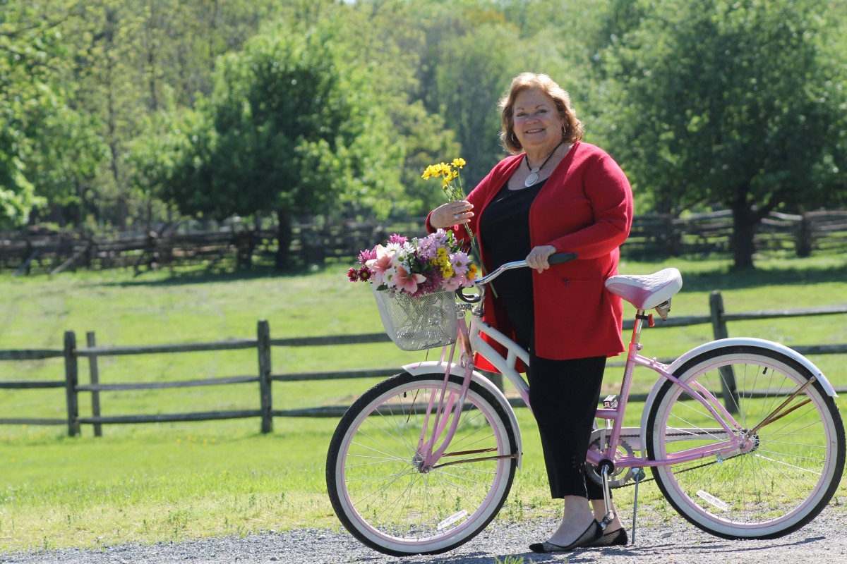 A woman stands beside a bicycle with a basket full of flowers in a scenic outdoor setting.