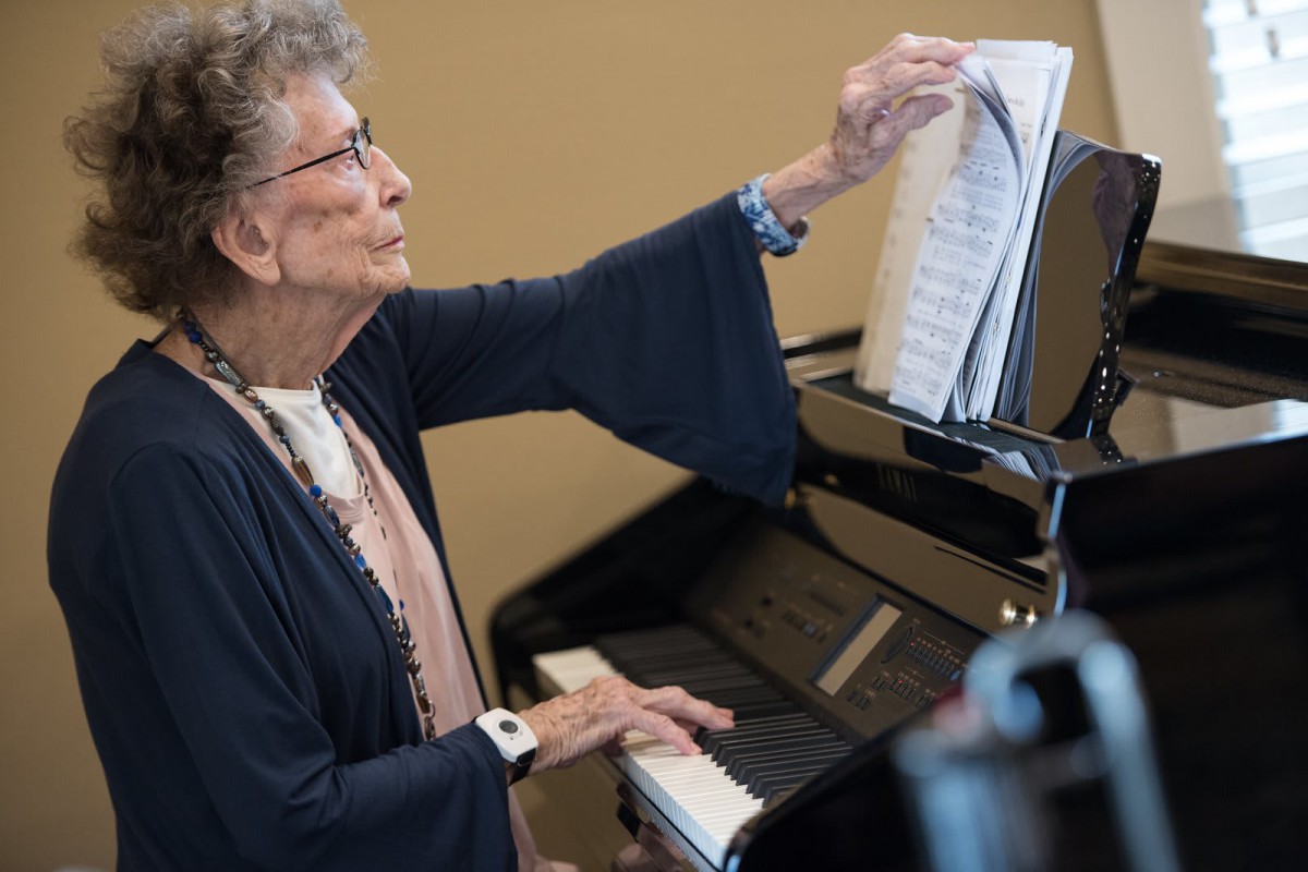 An elderly woman playing the piano while adjusting sheet music.