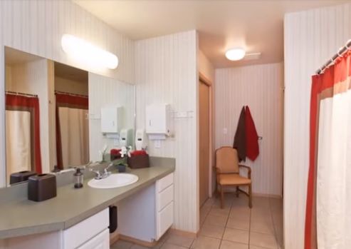 A bathroom with a vanity, sink, mirror, and light fixture. A towel hangs near a chair.