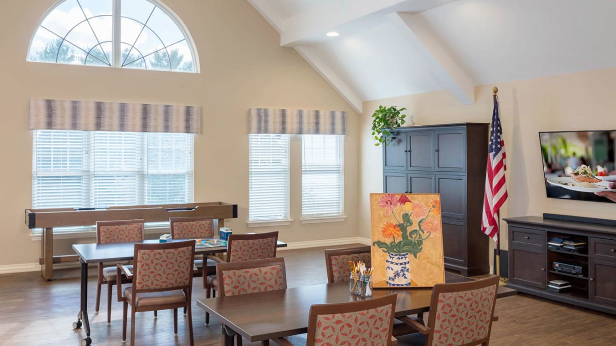 A cozy dining room with tables and chairs, featuring a colorful painting and large windows letting in natural light.