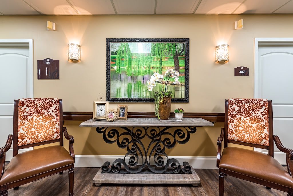 A warmly lit lobby area featuring a decorative table with two chairs, framed artwork, and plants.