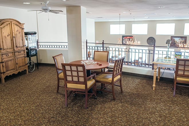 A cozy indoor area with a wooden table and chairs on a carpeted floor, featuring a ceiling fan and railing.