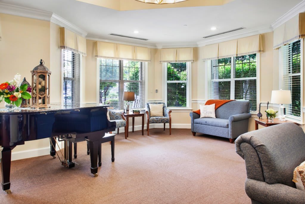 A cozy living room featuring a grand piano, sofas, chairs, and large windows allowing in plenty of natural light.
