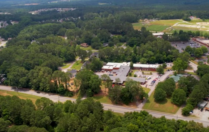 Aerial view of buildings surrounded by lush green trees and a road in a countryside setting.