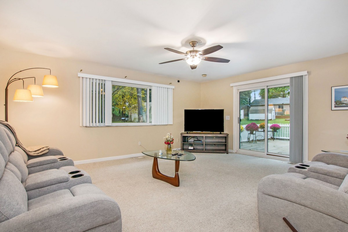 A spacious living room with a ceiling fan, TV, and modern glass table, featuring large windows and a sliding door leading outside.