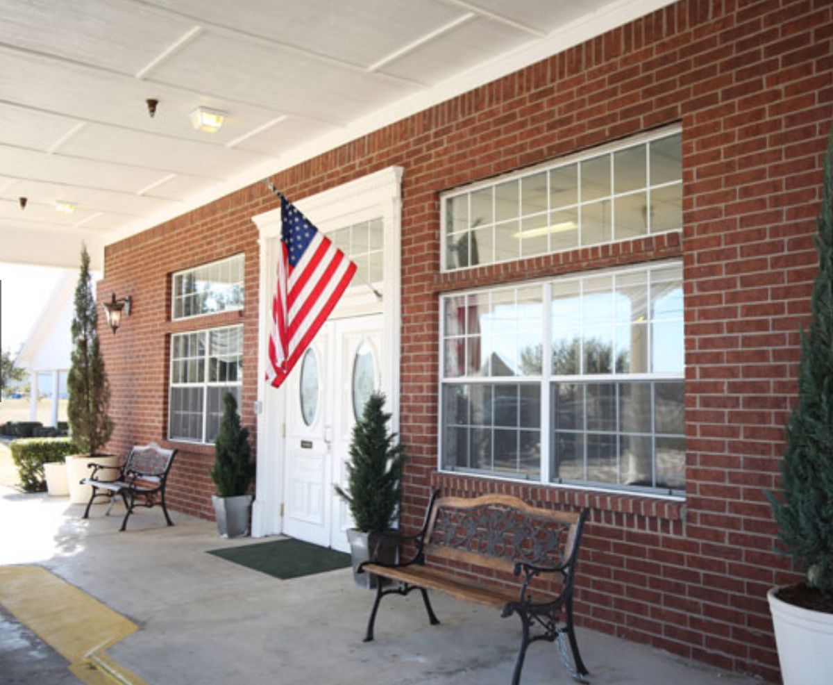 A brick building with an American flag, benches, and potted plants by the entrance.