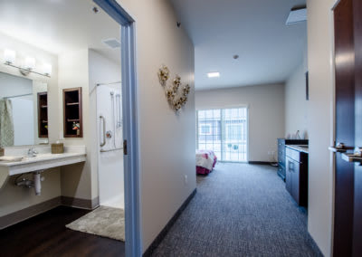 A modern interior featuring a hallway leading to a bedroom with a visible bathroom on the left and large windows in the background.