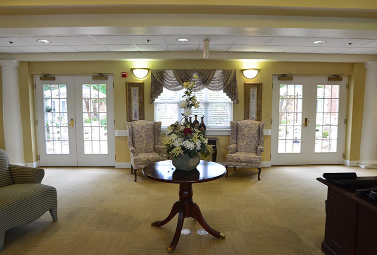 A cozy lobby area with a central table adorned with flowers, flanked by two chairs and surrounded by windows.