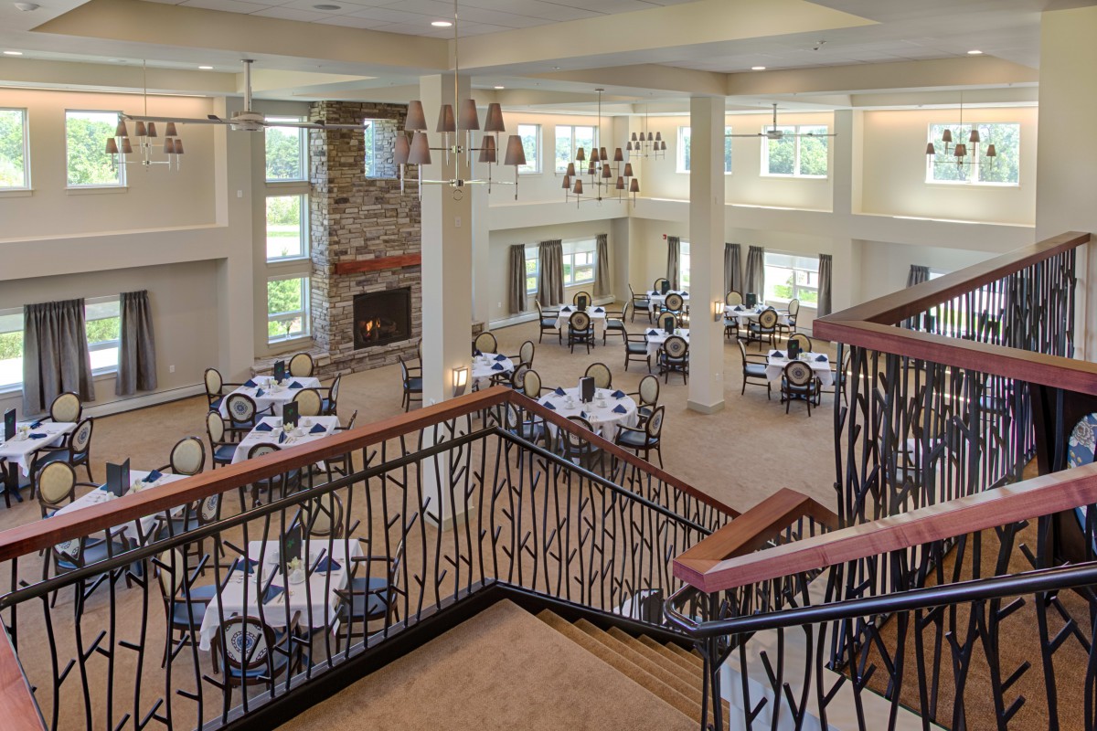 A spacious dining hall with elegant chandeliers and a grand staircase, featuring several tables set for guests.