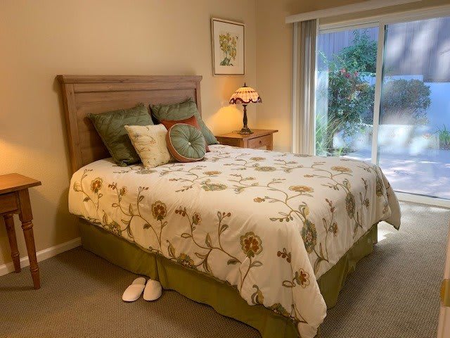 A cozy bedroom featuring a floral comforter on the bed, accent pillows, a bedside lamp on the nightstand, and a pair of slippers on the floor.