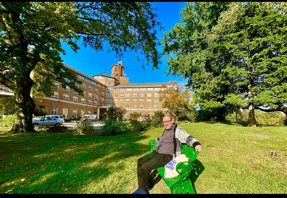 A person sitting on a green bench in a park with a large building in the background on a sunny day.