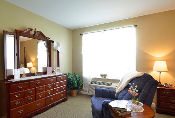 A cozy bedroom with a wooden dresser topped with a mirror, an upholstered armchair, and a large window allowing natural light.
