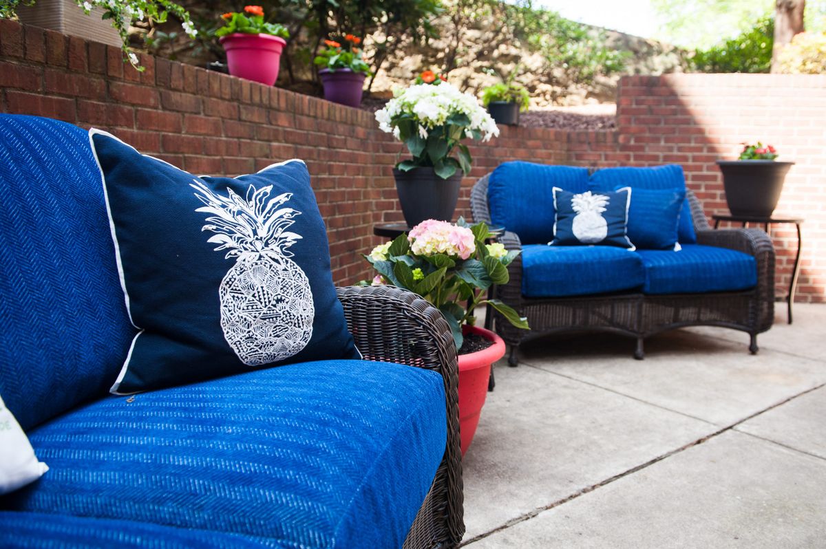 A cozy outdoor patio with wicker furniture featuring blue cushions and pineapple-themed decorative pillows, surrounded by potted plants against a brick wall.