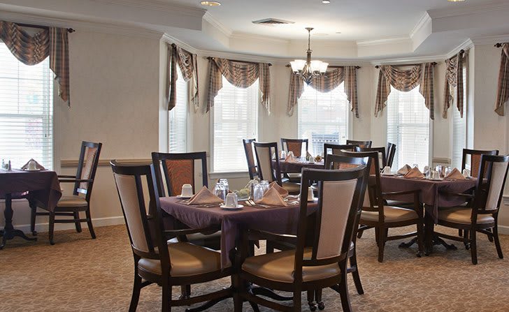 A well-lit dining area with elegantly set tables and chairs, surrounded by windows with curtains.