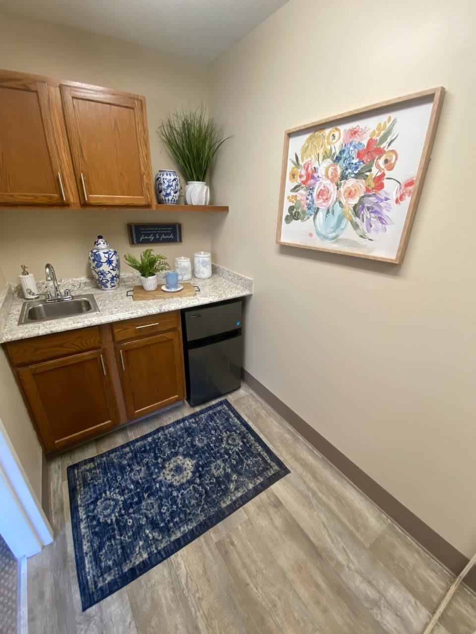 A small kitchenette with wooden cabinets, a mini fridge, and decorative elements such as vases and framed art.