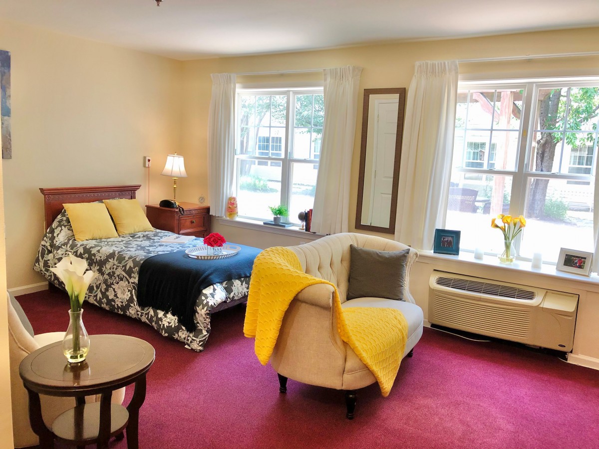 A cozy bedroom with a bed, chair, and large windows, featuring warm decor and red carpet.
