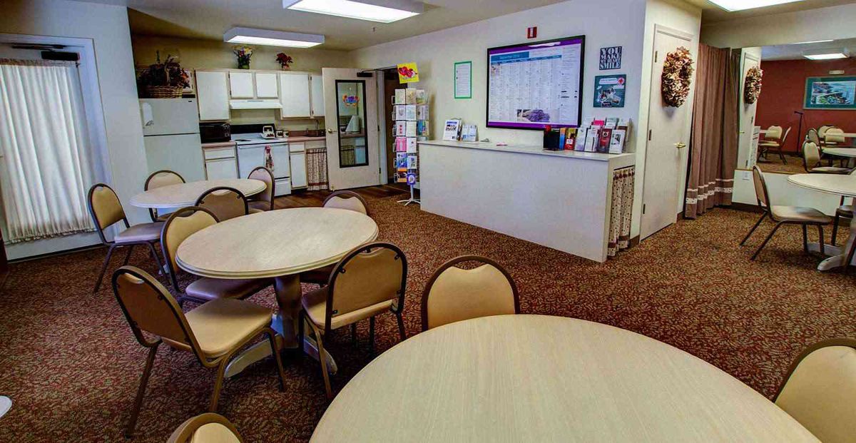 A community room with a kitchen and dining area featuring round tables and chairs on a carpeted floor.