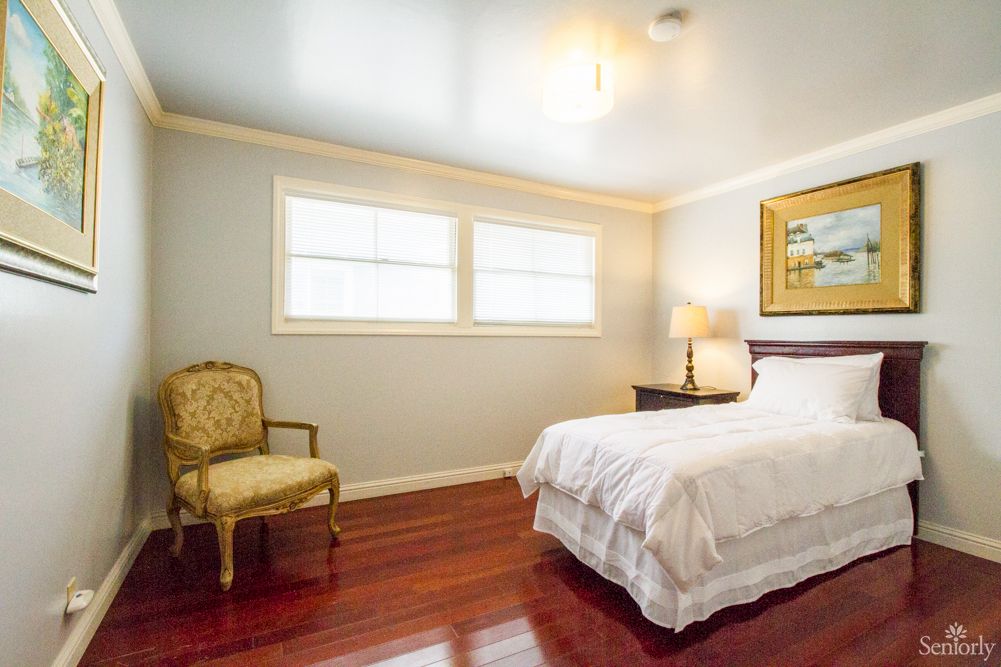 A cozy bedroom with a neatly made bed, an elegant chair, and warm wooden flooring.