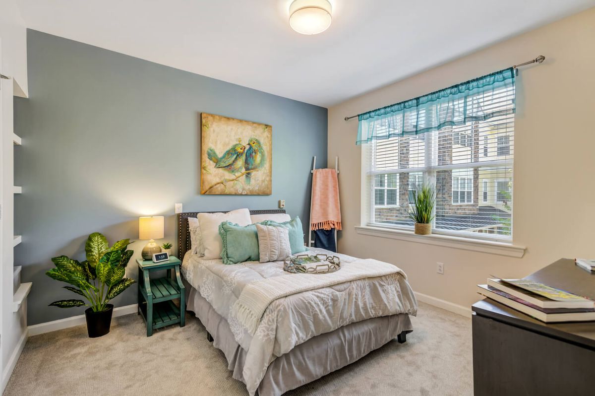 A modern and cozy bedroom with elegant decor, featuring a neatly made bed, artwork on the wall, and plants by the window.