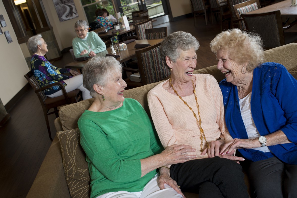 Three elderly women are sitting on a couch, laughing and enjoying each other's company in a cozy indoor setting.