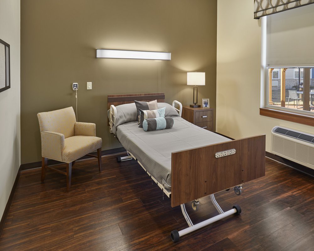 A modern hospital room with a single bed, chair, and warm lighting.
