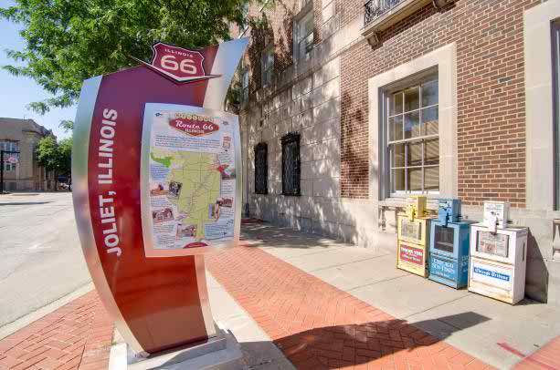 A Route 66 informational sign in Joliet, Illinois, displayed on a sidewalk next to a building.
