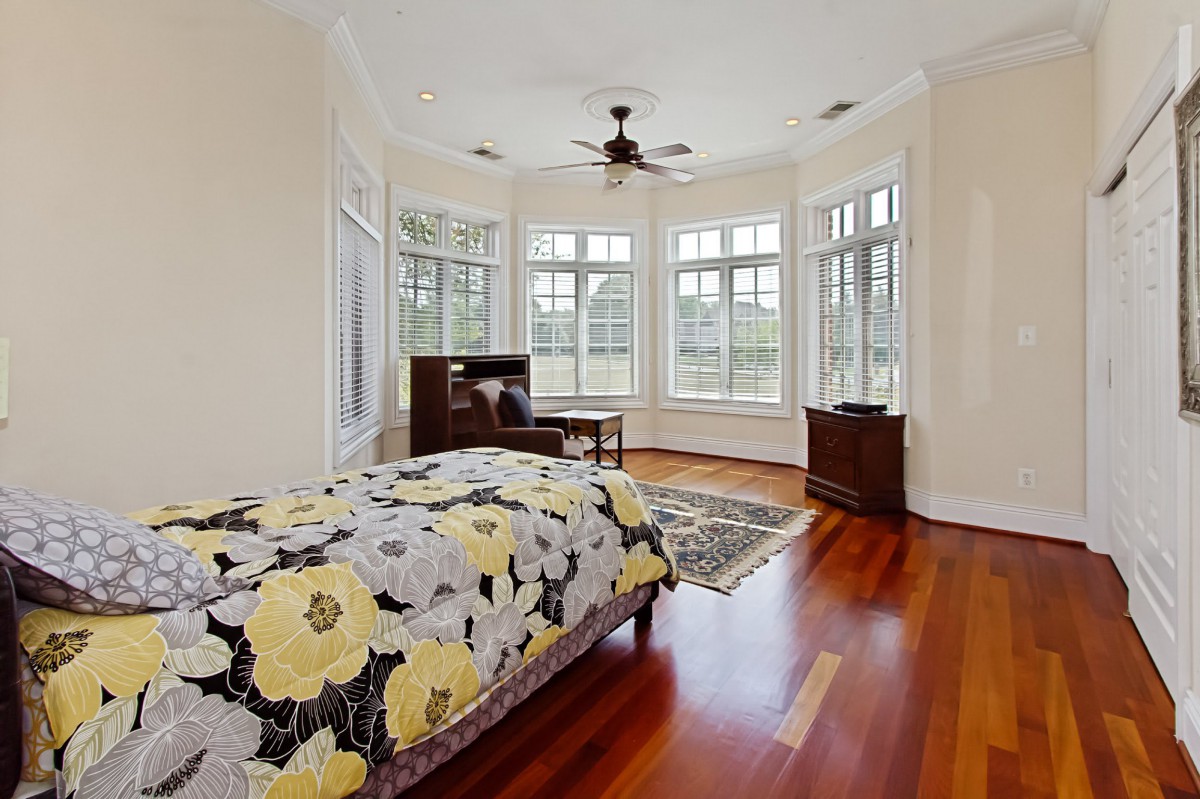 A spacious bedroom with wooden floors, large windows allowing natural light, and a floral bedspread.