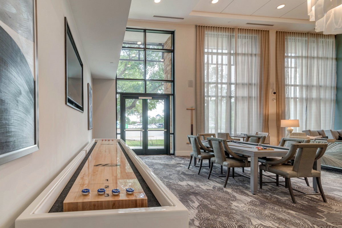 A modern living room featuring a shuffleboard table and a dining area with large windows.