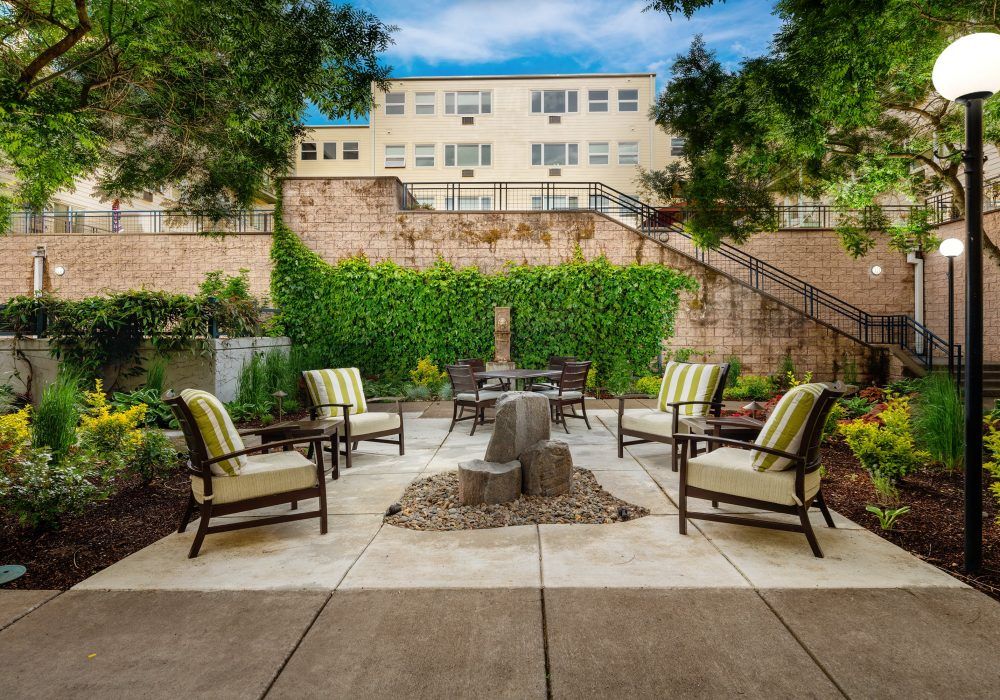 A cozy outdoor patio with chairs surrounding a fire pit, nestled in a courtyard with greenery and a building in the background.