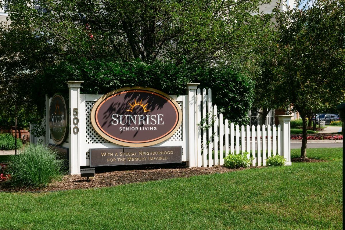 A welcoming sign for Sunrise Retirement, surrounded by greenery and a white picket fence.