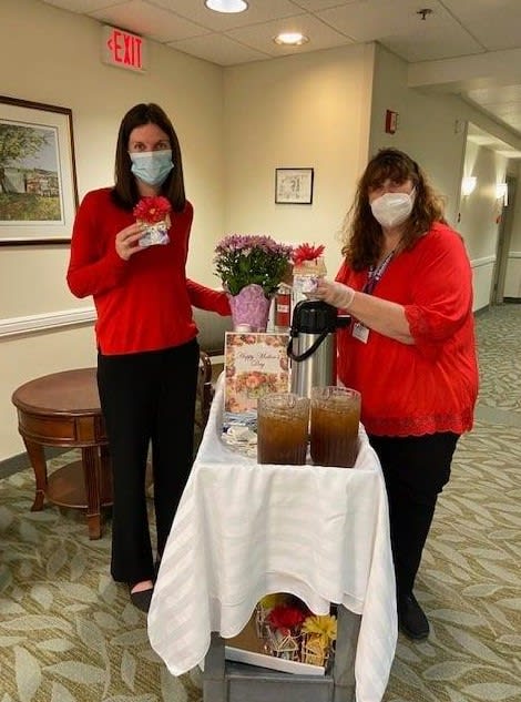 Two individuals wearing red clothing and masks are standing by a table with beverages and flowers.
