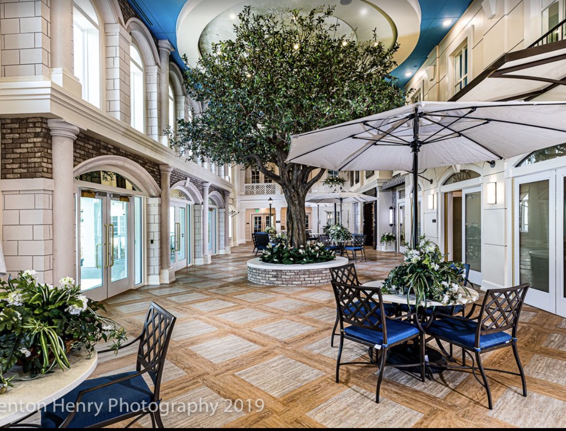 Luxurious indoor atrium featuring a large tree, elegant seating area with tables and umbrellas, surrounded by natural light.