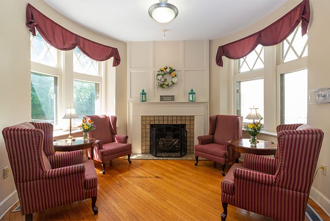 A cozy, traditionally decorated living room with a fireplace and comfortable armchairs, illuminated by natural light from large bay windows.