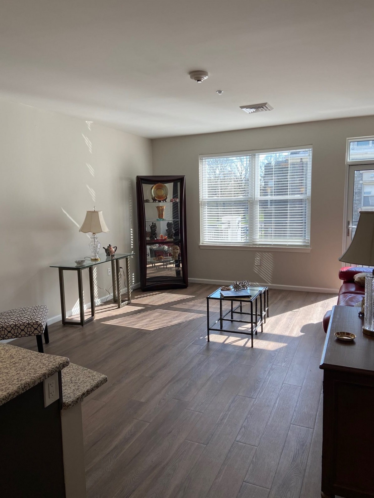 A bright living room with a wooden floor, featuring tables, lamps, and a cabinet near the window.