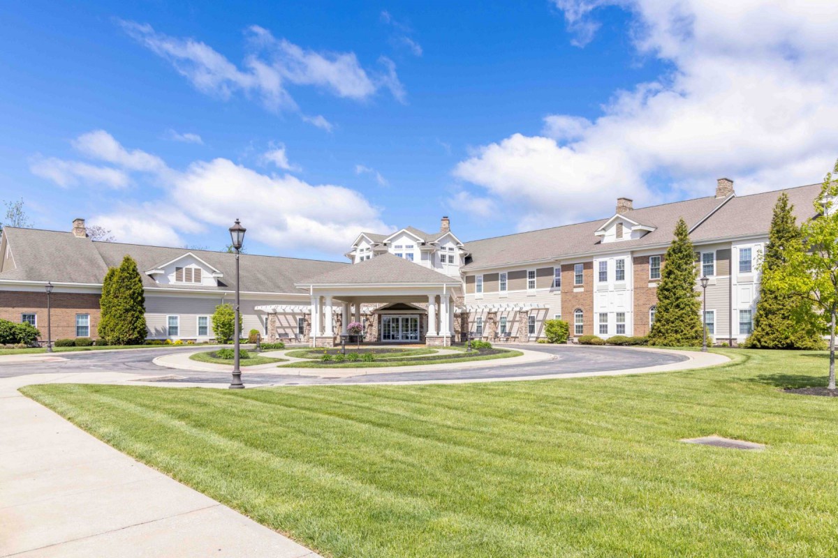A large, elegant building with a circular driveway and neatly trimmed grass under a bright blue sky with scattered clouds.