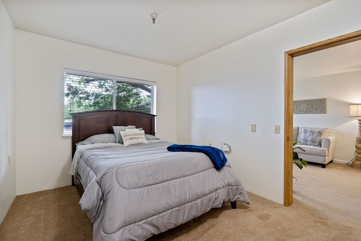 A cozy bedroom featuring a neatly made bed with a wooden headboard, complemented by a blue blanket and pillows. The room is well-lit by natural light coming through the window.