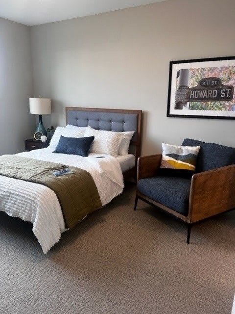 A modern bedroom featuring a cozy bed with white and olive bedding, a stylish armchair, and framed artwork on the wall.