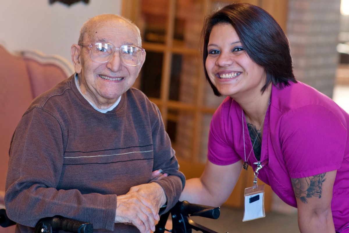 An elderly man in a wheelchair smiling alongside a caregiver indoors.