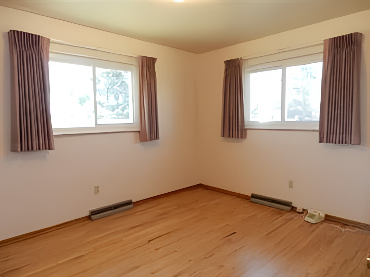 An empty room with hardwood floors, two windows with curtains, and baseboard heaters.