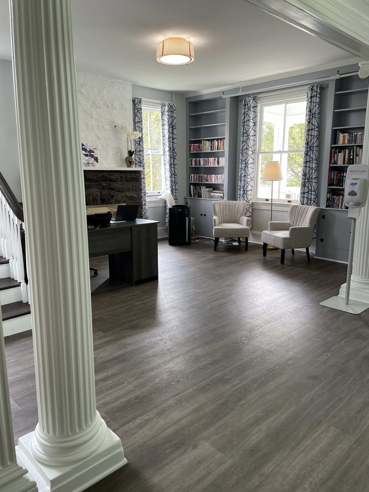 A spacious living room with hardwood floors, featuring elegant pillars, a desk area, and large windows flanked by bookshelves.
