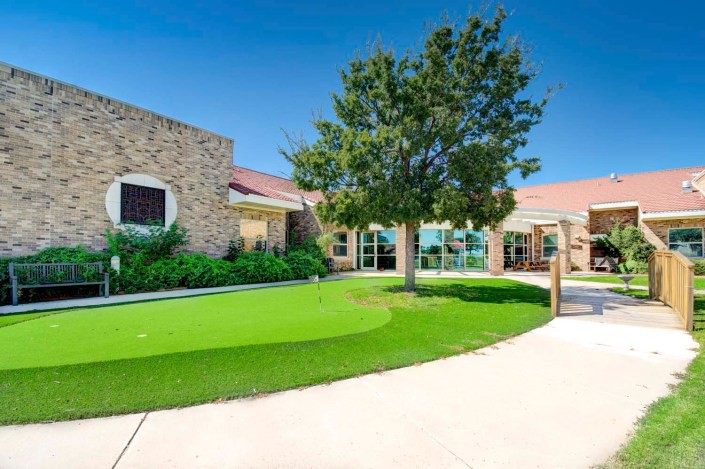 A modern building with stone accents surrounded by a well-maintained lawn and a large tree under a bright blue sky.