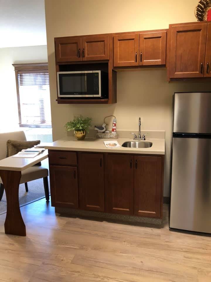 A compact kitchenette with wooden cabinetry, featuring a microwave, stainless steel fridge, and sink.