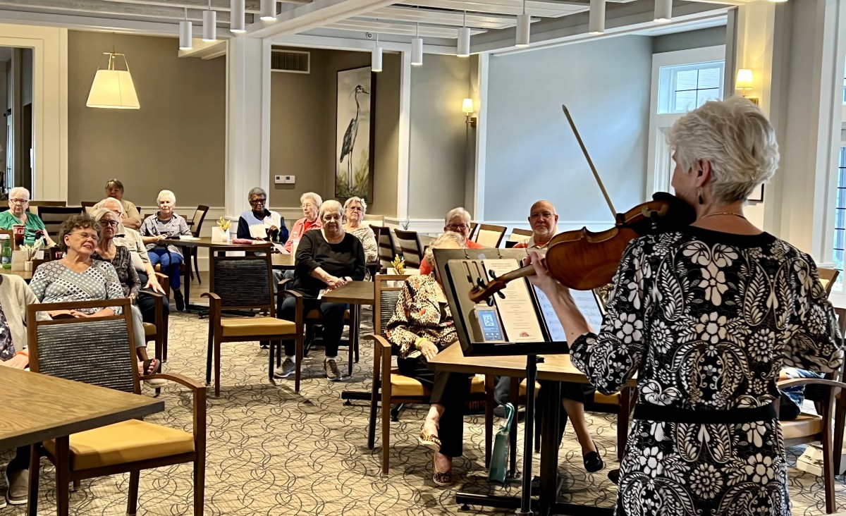 A violinist performs for an audience seated in a bright indoor venue.