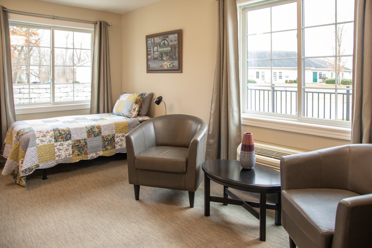 A cozy room with two armchairs, a small table, a bed, and large windows.